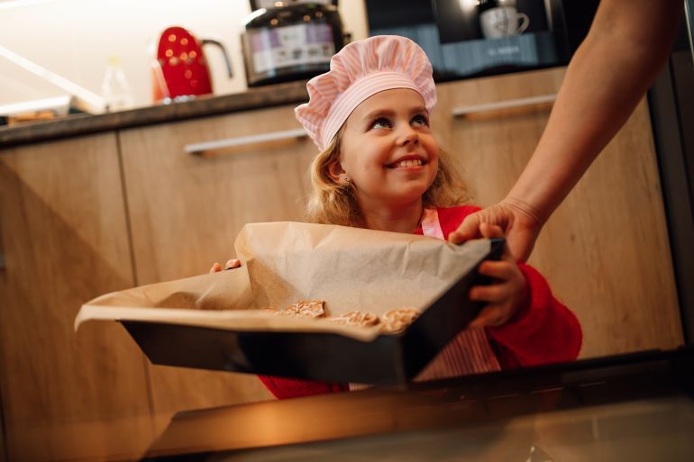 A cute young girl, dressed in a chef's hat and apron, carefully takes a baking pan of freshly baked goods out of the oven, showing a joyful expression. The image is perfect to illustrate the theme of cooking, baking, childhood and family activities.