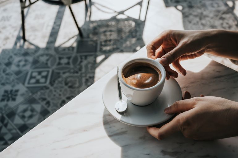 Close up of woman's hand holding a cup of coffee, drinking coffee in outdoor cafe against beautiful sunlight, having a relaxing moment. Enjoying life's simple pleasures