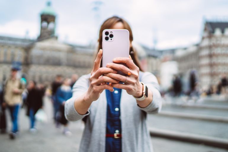A young female traveller using her smart phone to capture a moment at a bustling historic square in Amsterdam. The focus is on her hands holding the device, with the vibrant city life and architectural beauty in the background. Concept of urban exploration, solo travel and vacation.