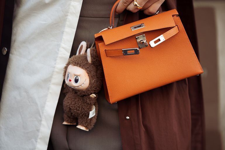 PARIS, FRANCE - MARCH 08: A guest wears brown coat, white blouse, orange Hermes bag with a bag charm brown Labubu monster, grey pants outside the Hermes fashion show during the Womenswear Fall/Winter 2025/2026 as part of Paris Fashion Week on March 08, 2025 in Paris, France. (Photo by Raimonda Kulikauskiene/Getty Images)