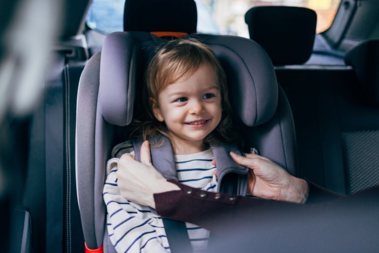 Portrait of a cute girl excited while sitting in a car safety seat. Her anonymous mother helping her little daughter to buckle and fasten seatbelt properly to stay safe while driving.